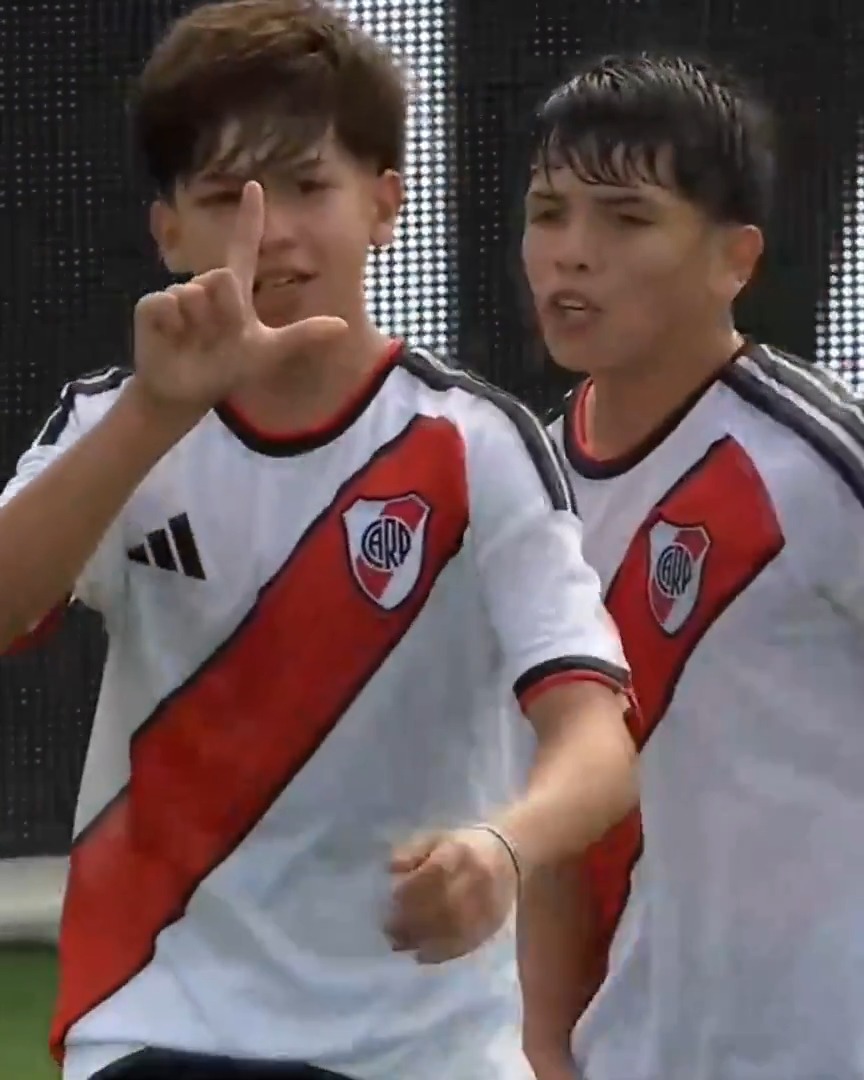 Two young soccer players in white River Plate jerseys with a red diagonal sash are shown on a pitch, with the player on the left making a hand gesture toward his eye.