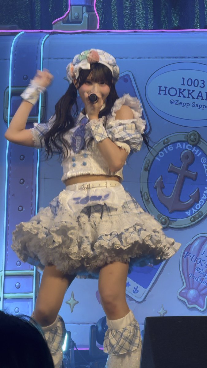 A performer in a frilly white and blue plaid outfit sings into a microphone on stage, with a backdrop featuring suitcase graphics and location names like "HOKKAIDO" and "AICHI".