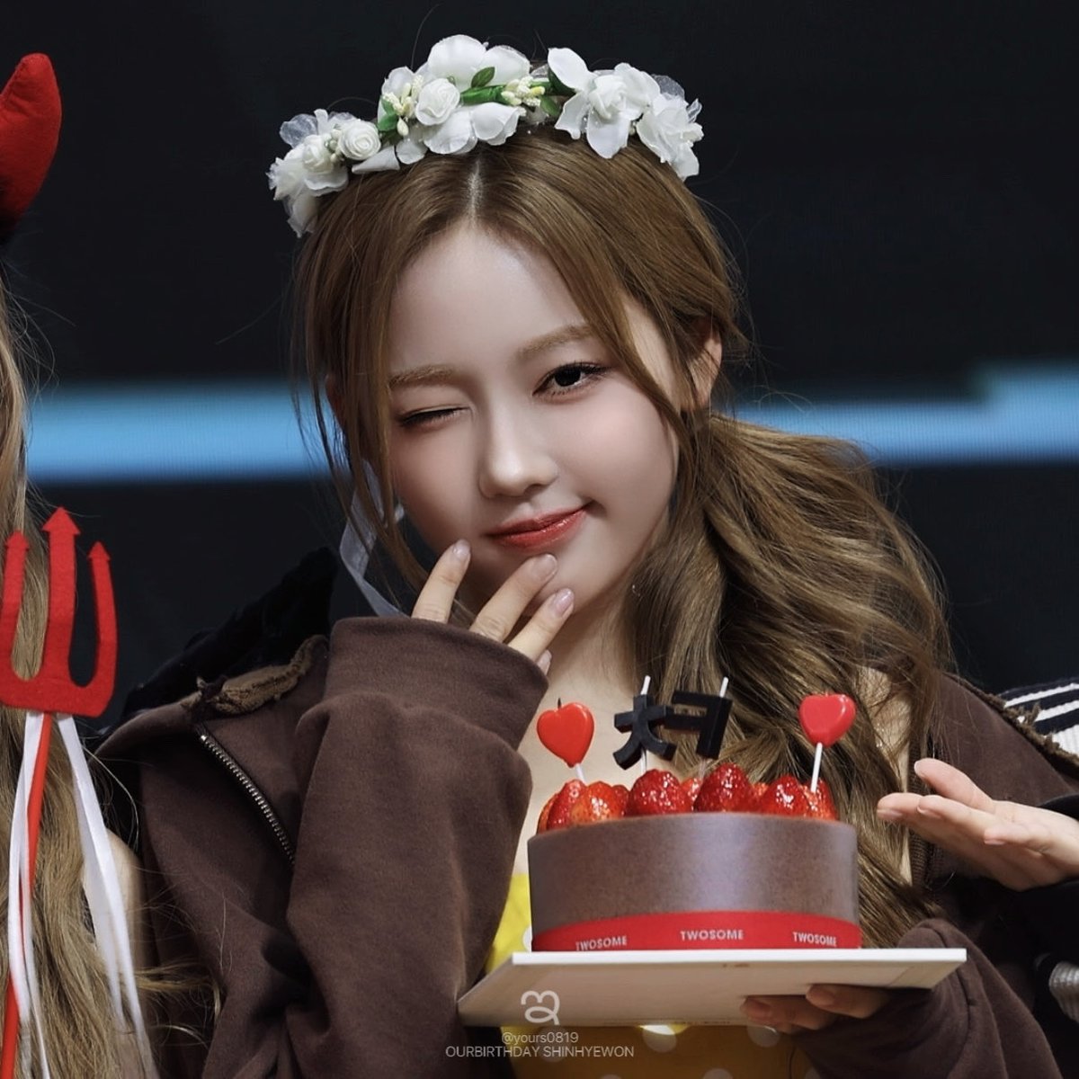 A young woman with long brown hair and a white flower crown winks while holding a strawberry cake with red heart toppers; the cake wrapper reads 'TWOSOME' and the watermark says 'OURBIRTHDAYSHINHYEWON'.