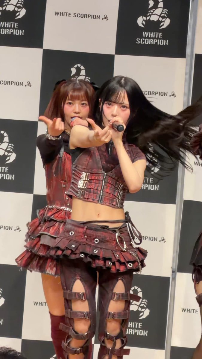 Two young women wearing matching red and black gothic-style outfits perform on stage with microphones, posing in front of a checkered backdrop with 'WHITE SCORPION' logos.