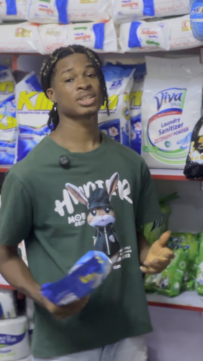 A young man with braided hair, wearing a green 'Hunter Mob Grip' t-shirt, stands in front of store shelves stocked with laundry detergent and holds a blue package.