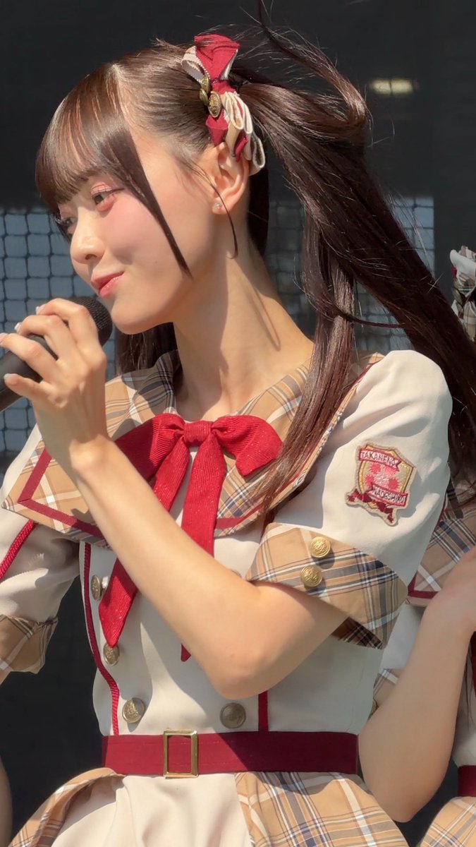 A young woman performs on stage wearing a plaid and white dress with a red bow, holding a microphone with her hair in pigtails tied with red bows.