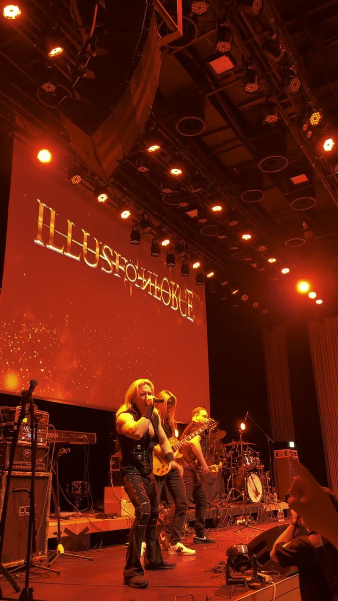 A band performs on stage bathed in red lighting under a large screen displaying the word 'ILLUSIONOGE' in gold lettering, with a singer at the microphone and guitarists playing.