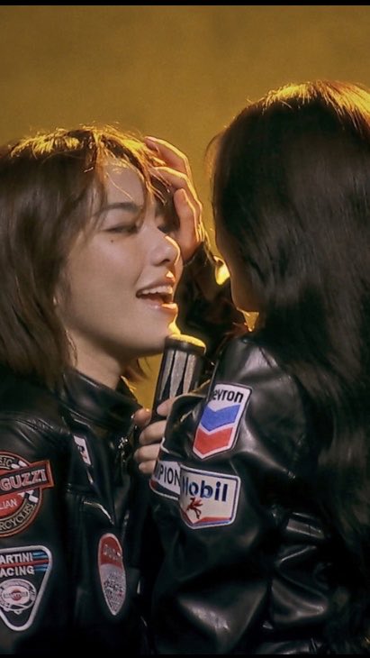 A close-up of two people wearing black leather jackets with racing patches including 'Guzzi', 'Mobil', and 'Martin Racing'; one person is smiling warmly at the other.