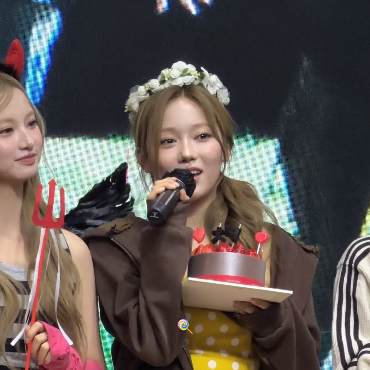A performer wearing a flower crown speaks into a microphone while holding a chocolate cake decorated with hearts, standing next to another person holding a red trident prop.