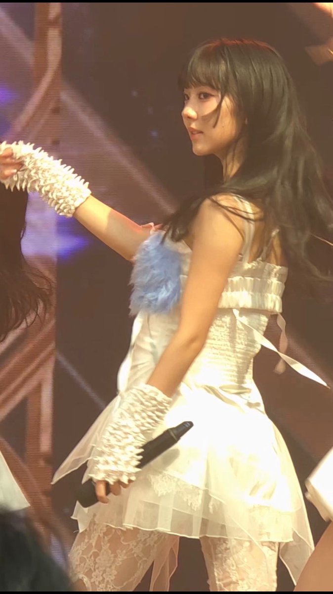 A young woman performer with long dark hair on stage, wearing a white ruffled dress with lace details and long white fingerless gloves, with a blue fluffy accent on her chest, holding a microphone.
