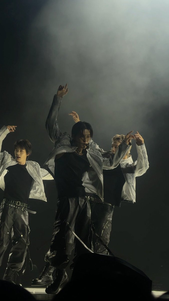 Three performers in black and silver outfits pose dramatically on a dimly lit stage with one raising their arm high, suggesting a live concert performance.