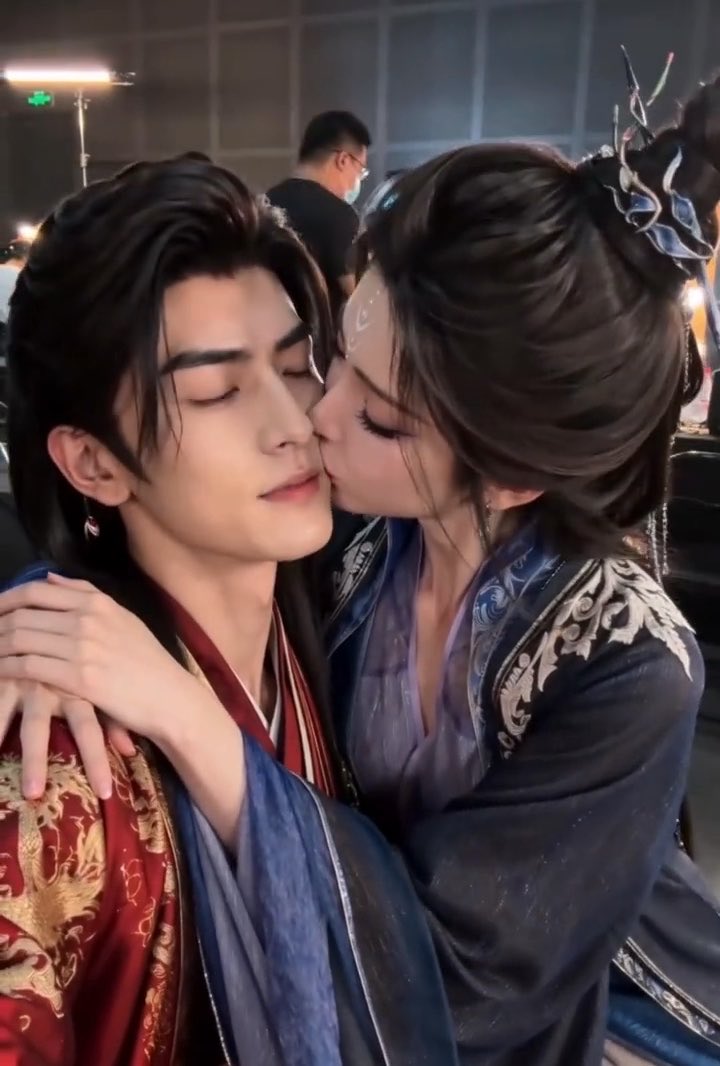 Two people in ornate traditional Chinese costume, with the woman on the right kissing the cheek of the man on the left who has his eyes closed, seemingly a behind-the-scenes shot from a drama production.