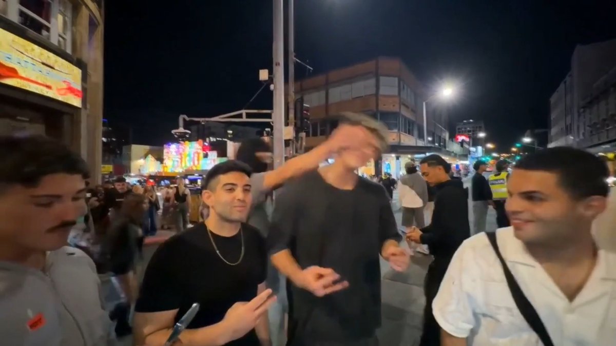 A nighttime street scene showing several young men, including one blurred figure in the center with gray hair and a gray t-shirt, surrounded by others smiling, with police officers and illuminated signs visible in the busy background.