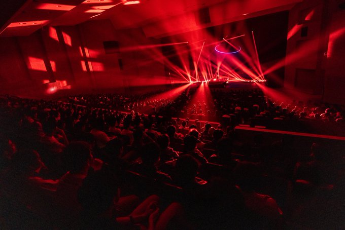 A concert venue is bathed in red lighting, with beams shining from the stage over a seated audience.