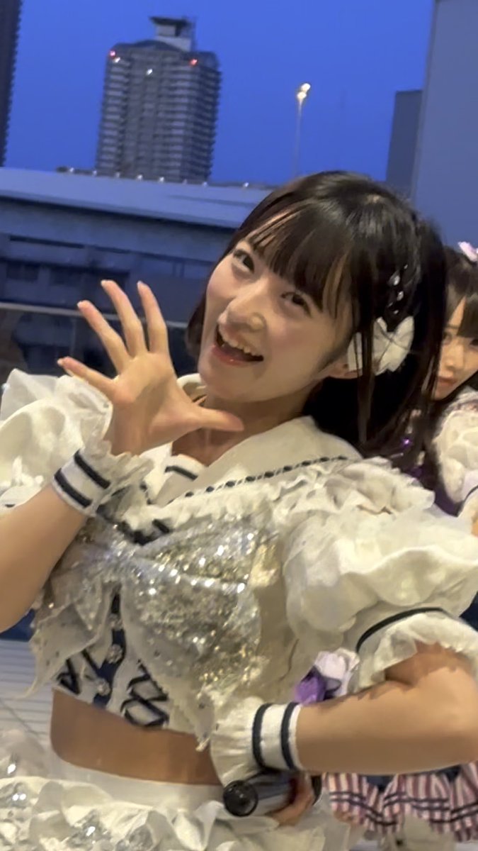 A young woman in a frilly white and blue outfit waves at the camera, with a tall building visible in the twilight background.