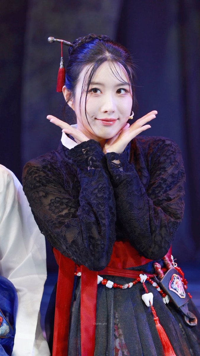 A young woman in traditional Korean hanbok attire with a black lace top and red accents, posing playfully with her hands framing her face.
