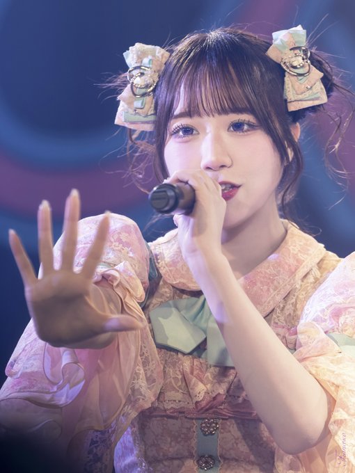 A young woman with hair bows in a pink frilly dress sings into a microphone while reaching her hand toward the camera.