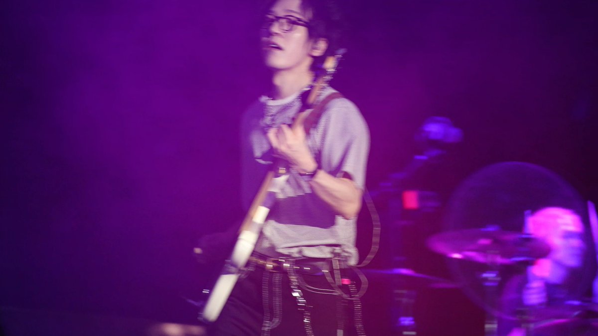 A blurry purple-lit concert photo of a bespectacled musician playing guitar with a drummer partially visible in the background.