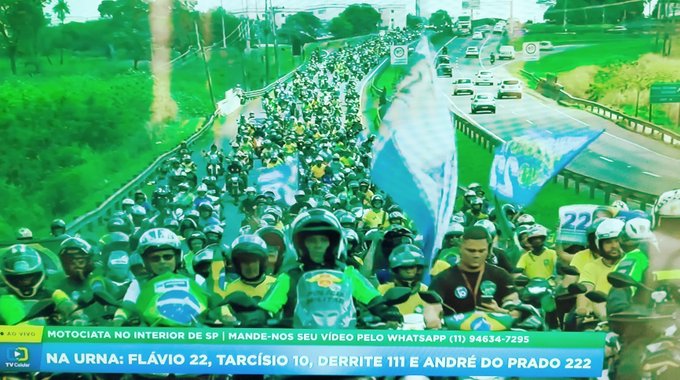 A news broadcast showing a massive motorcycle rally on a highway, with a ticker reading 'MOTOCIATA NO INTERIOR DE SP | MANDE-NOS SEU VÍDEO PELO WHATSAPP (11) 94634-7295' and 'NA URNA: FLÁVIO 22, TARCISIO 10, DERRITE 111 E ANDRÉ DO PRADO 222'.