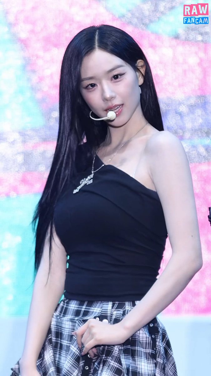 A fancam screenshot of a female performer on stage with long black hair, a headset microphone, and a black asymmetrical top over a plaid skirt, against a pink and blue digital backdrop.