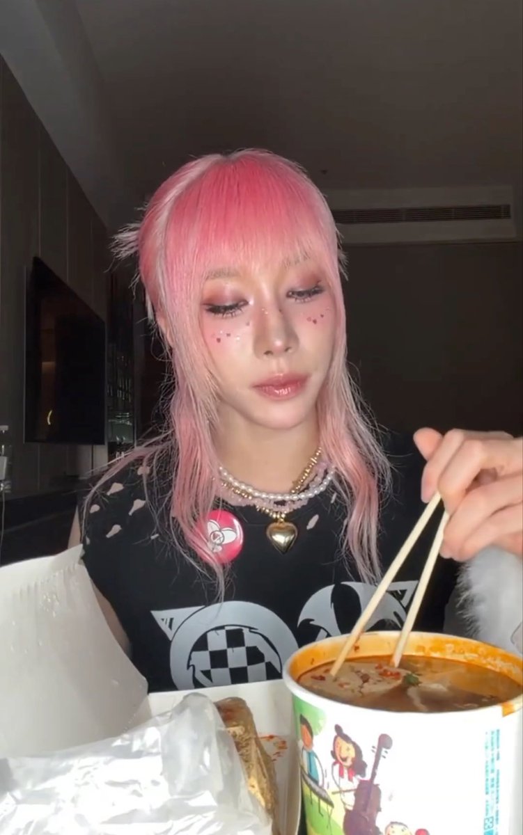 A young woman with pink hair and small heart details on her cheeks eats from a cup of food with chopsticks, wearing a black graphic t-shirt.