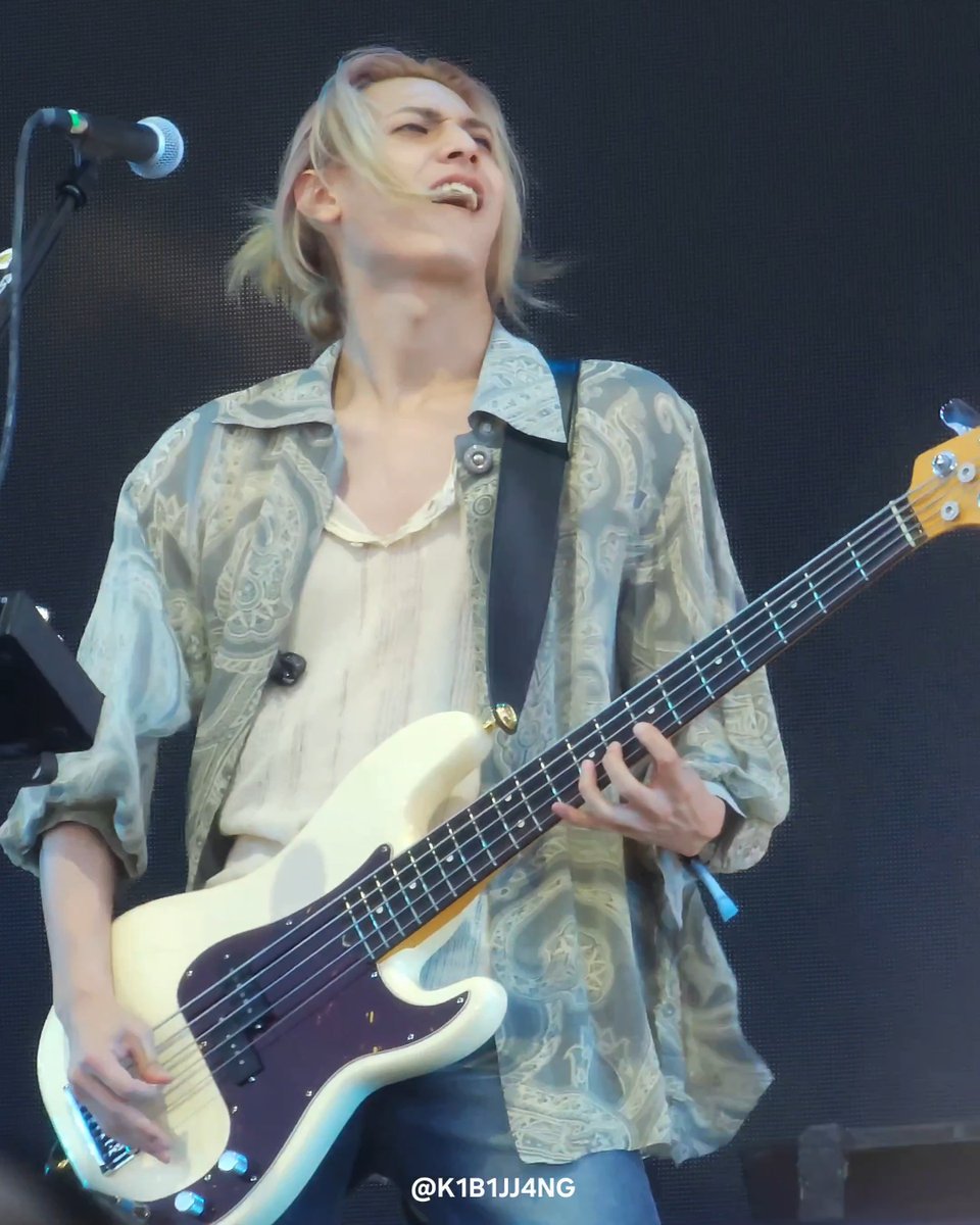 A blonde musician passionately plays a white bass guitar on a dark stage, wearing a sheer patterned shirt, with a watermark '@K1B1JJ4NG' in the corner.