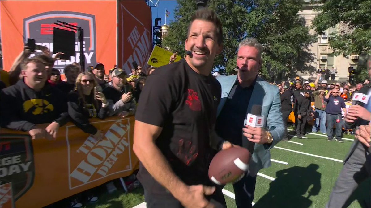 ESPN College GameDay host Drew Carter interviews former Iowa Hawkeyes tight end Dallas Clark, who smiles while holding a football, with fans in black and gold cheering behind a Home Depot-sponsored barricade. The scene captures the moment after Clark successfully threw a football through the uprights to win $150,000 for a fan and $150,000 for the hospital.
