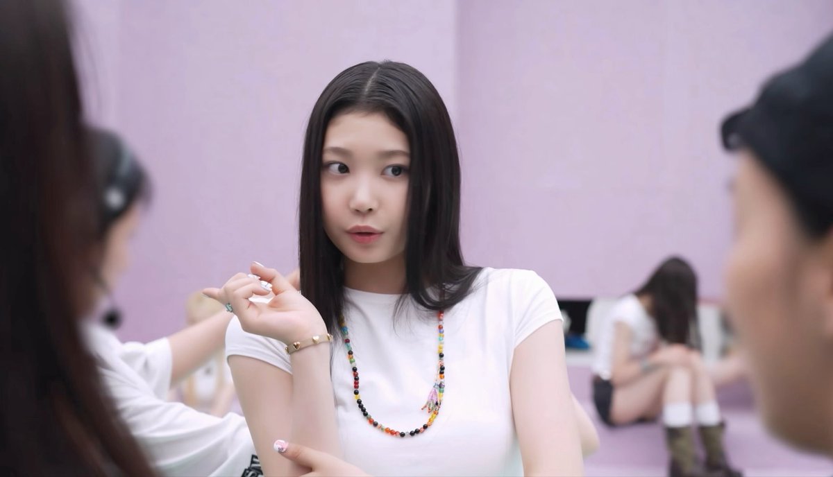 A woman with long dark hair wearing a white t-shirt and a colorful beaded necklace sits and looks to the side in a room with other people.