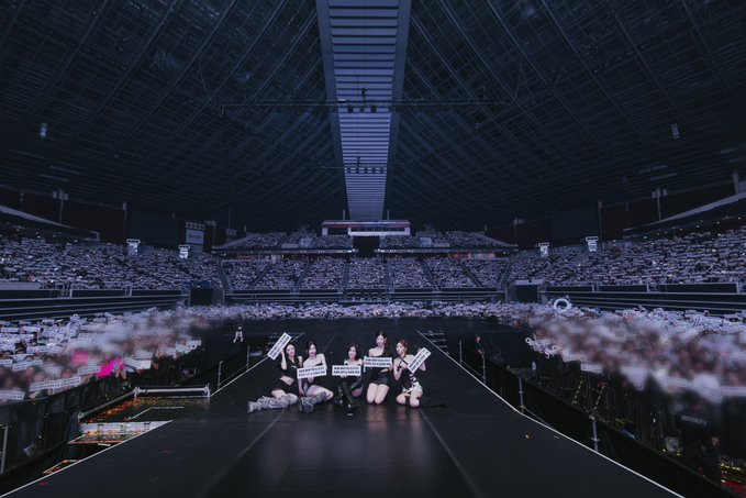 A wide shot of a large arena where a K-pop group performs on stage holding signs, with a massive crowd of fans holding up white balloons in the darkened stadium.