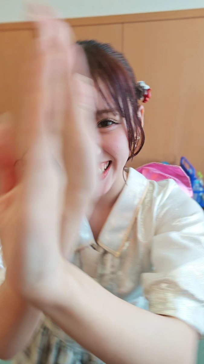 A young woman with reddish-brown hair smiles brightly while clapping her hands, captured in a candid, slightly blurry motion shot wearing a light-colored blouse.