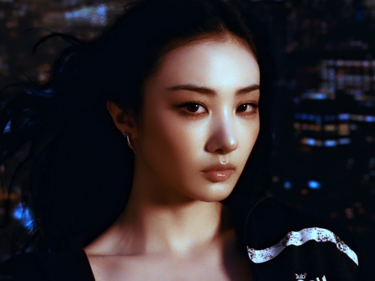 A stylized portrait of a young woman with dark hair and dramatic makeup, with city lights blurred in the background, appearing to be a digital artwork or heavily edited photo.