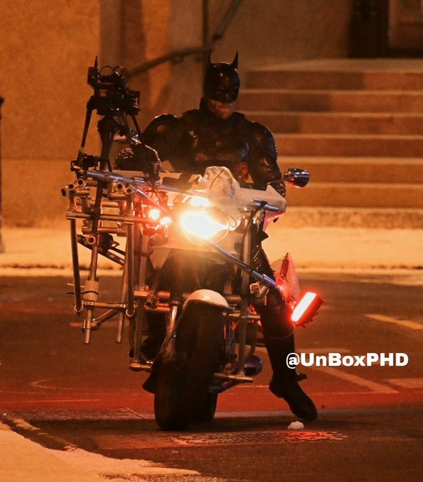 A nighttime behind-the-scenes photo shows someone in a Batman costume on a motorcycle with elaborate camera rigging attached, likely filming a chase scene for The Batman Part II. The watermark "@UnBoxPHD" is visible in the lower right, and snow-dusted steps appear in the background.