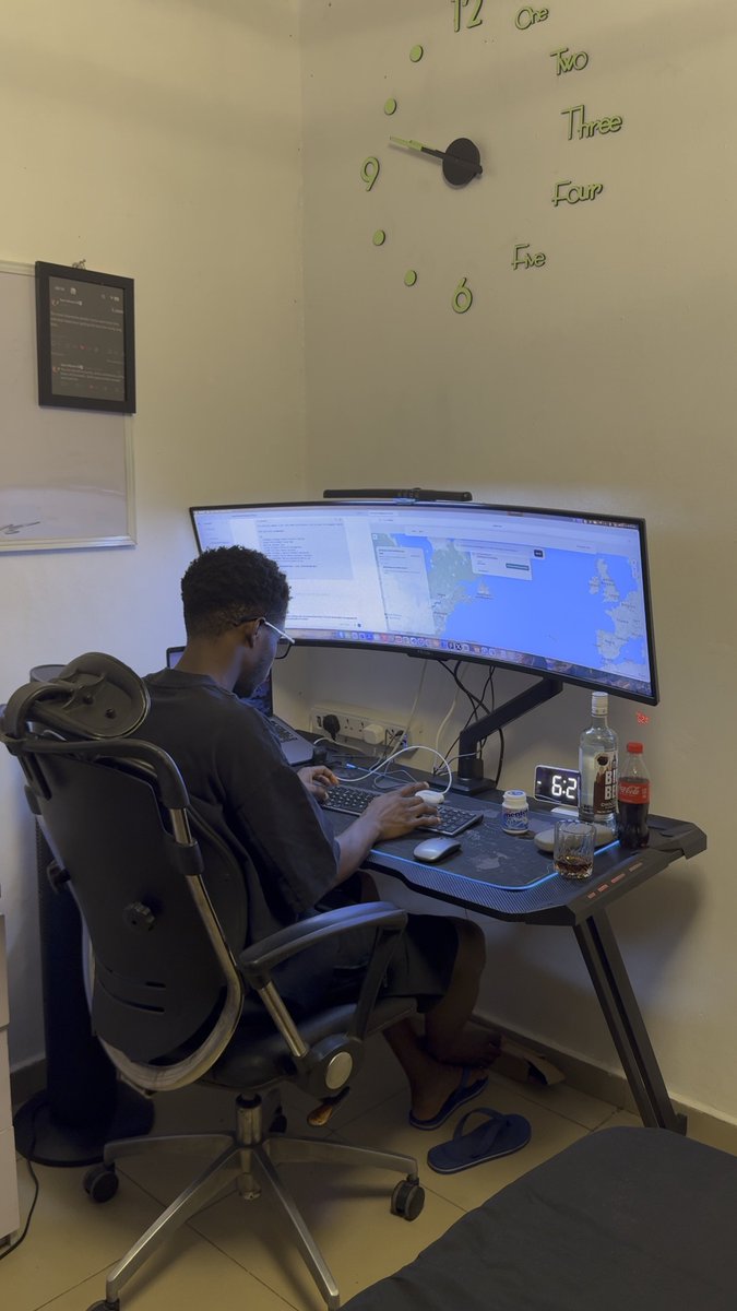 A young man works at a desk with an ultrawide curved monitor displaying maps and documents, with a wall clock above spelling numbers in words and a digital clock reading 6:21.