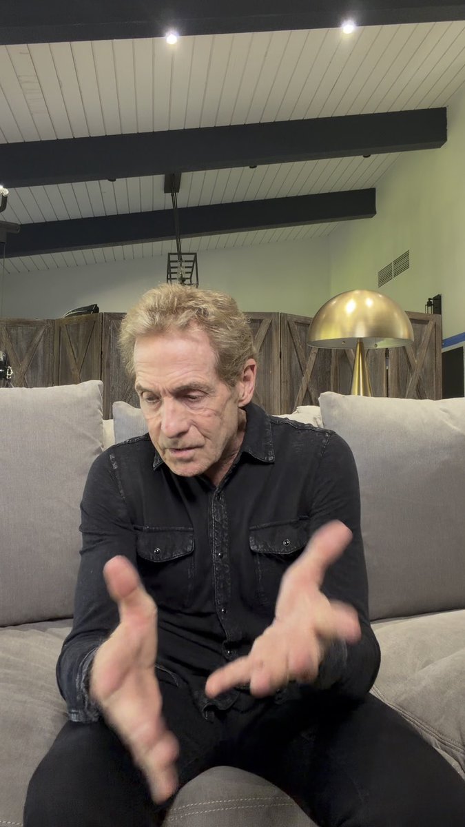 A gray-haired man wearing a black button-up shirt sits on a gray couch, gesturing expressively with both hands while speaking in a warmly lit room with wood-beamed ceilings. The image reflects the introspective tone of his Club Shay Shay interview with Shannon Sharpe, capturing a candid moment of reflection from the conversation.