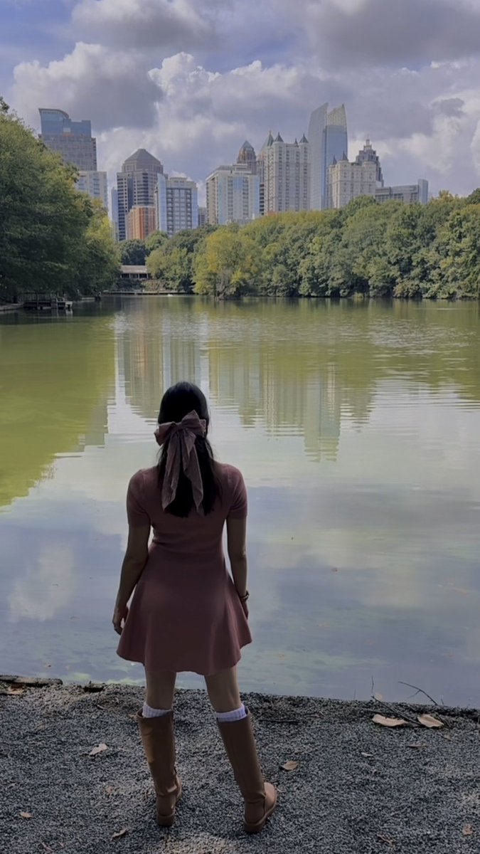 A woman in a pink dress with a hair bow stands with her back to the camera, looking out over a green lake toward a city skyline under a partly cloudy sky.