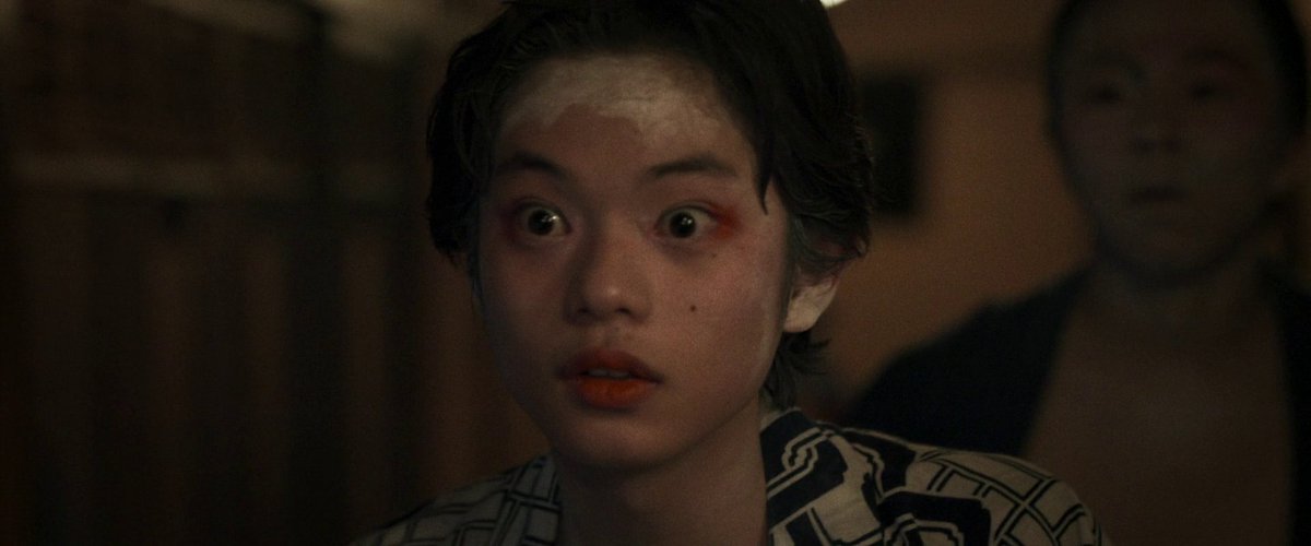 A young person with wide, startled eyes and an orange tint on their lips stares forward in a dimly lit room, with a blurred figure visible in the background. The image appears to be a still from the 2025 Japanese film "Kokuho," featuring actor Soya Kurokawa.