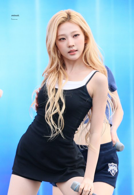 A performance photo of a blonde K-pop idol (credited to 'minuti.') wearing a black dress over a white top, holding a microphone on stage with a blue background.