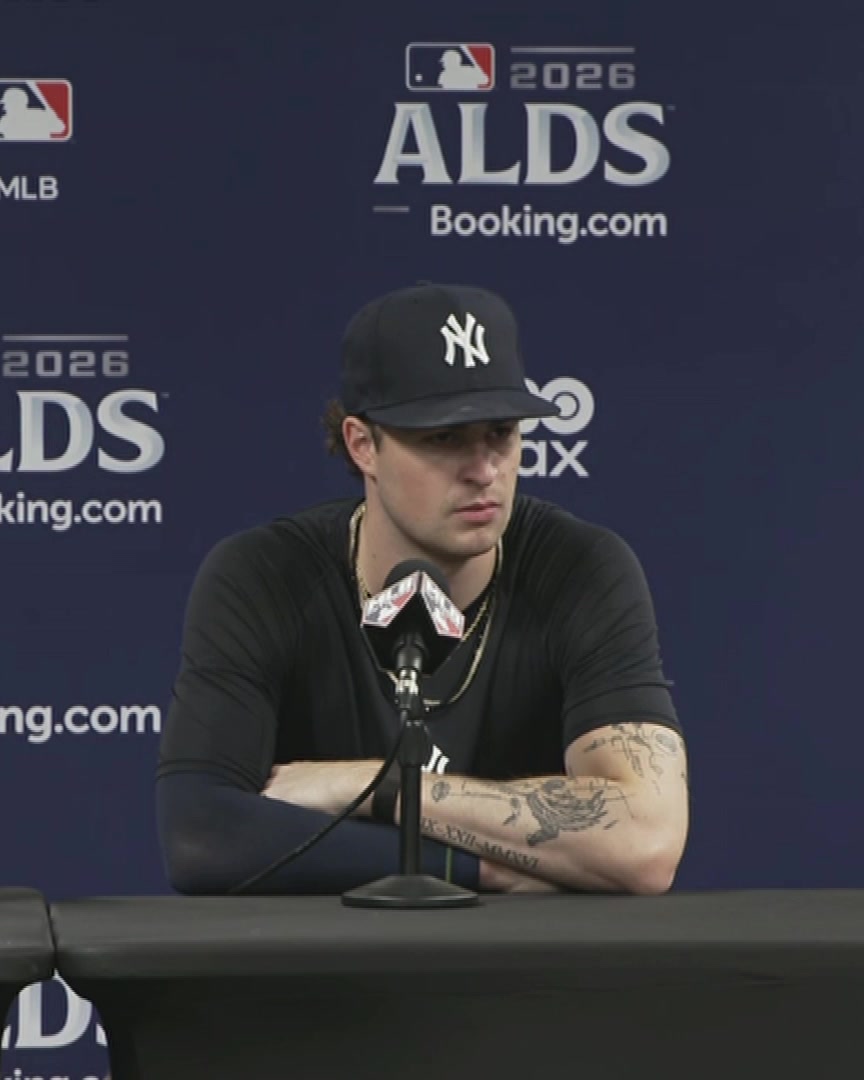 A player wearing a New York Yankees cap sits at a press conference table during the 2026 ALDS, with a backdrop featuring MLB and Booking.com logos, as he addresses reporters about the anticipated Yankee-heavy crowd at Tropicana Field.