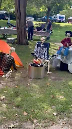 A Halloween-themed campground scene shows skeletons dressed in plaid shirts and skirts seated around a fire pit with a real person visible in the background near a picnic table.