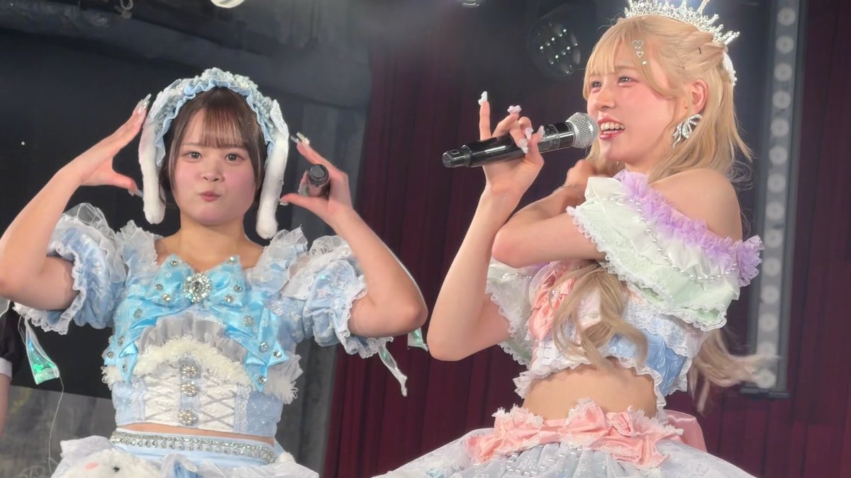 Two performers in elaborate, pastel-colored frilly idol costumes are on stage; one holds a microphone while the other makes a playful gesture with her hands.