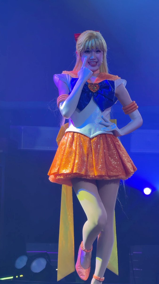 A performer dressed as Sailor Venus from Sailor Moon poses on stage in an orange and blue sailor uniform with a sparkling orange skirt, smiling and pointing upward against a blue-lit background.