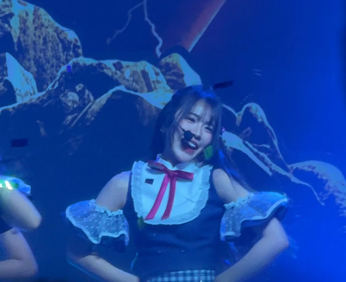 A performer in a blue outfit featuring a large white collar with a pink bow and a head microphone is captured mid-performance on a blue-lit stage.