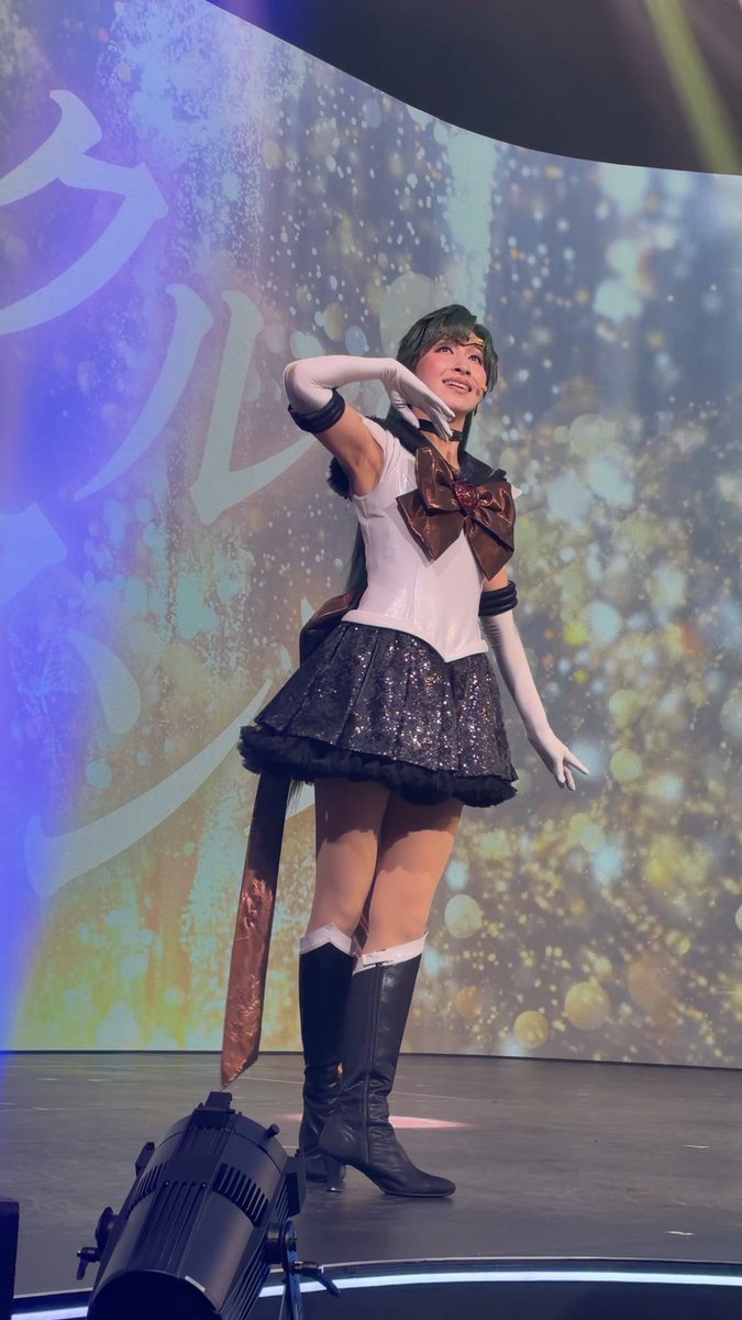 A performer on stage poses in a Sailor Moon-inspired costume featuring a white top, dark pleated skirt, large bronze bow, white gloves, and black knee-high boots.