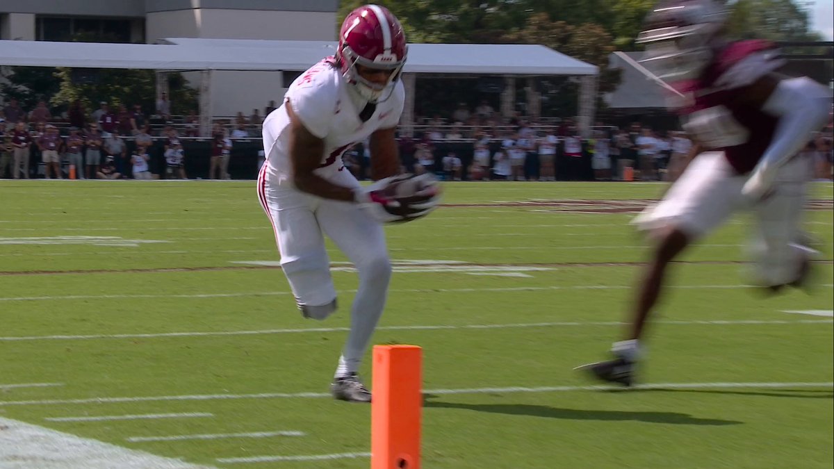 A Crimson Tide football player, likely Ryan Coleman-Williams, sprints toward the end zone pylon with the ball secured in his arms, having just scored a touchdown to give Alabama the early lead. The orange pylon in the foreground marks the goal line as a defender trails behind in pursuit.