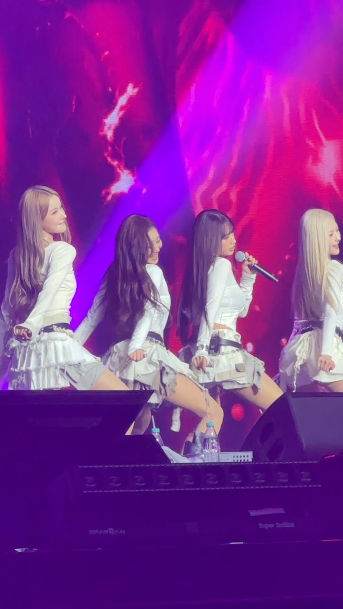 A concert photo of four female K-pop performers in white outfits performing on stage with purple and red lighting.