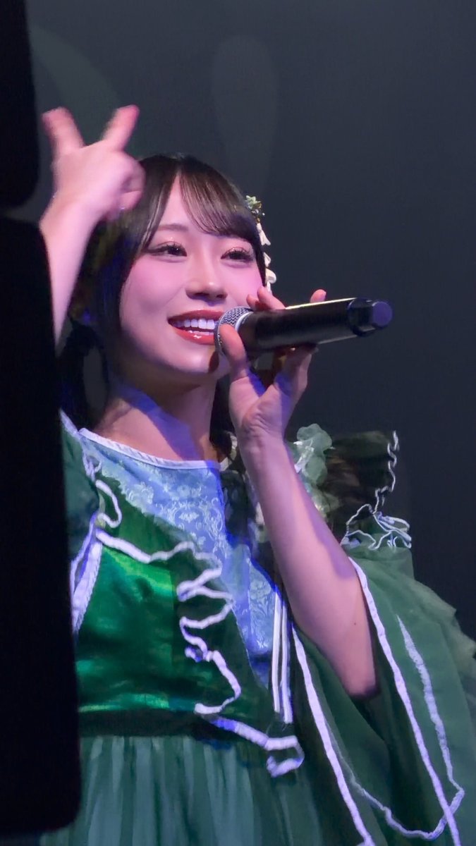 A young woman with dark hair smiles brightly while singing into a microphone on stage, wearing a green dress with white ribbon details.