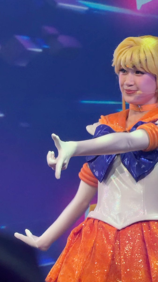 A performer dressed as Sailor Venus in an orange and white costume with a blonde wig and tiara poses on stage with a headset microphone.