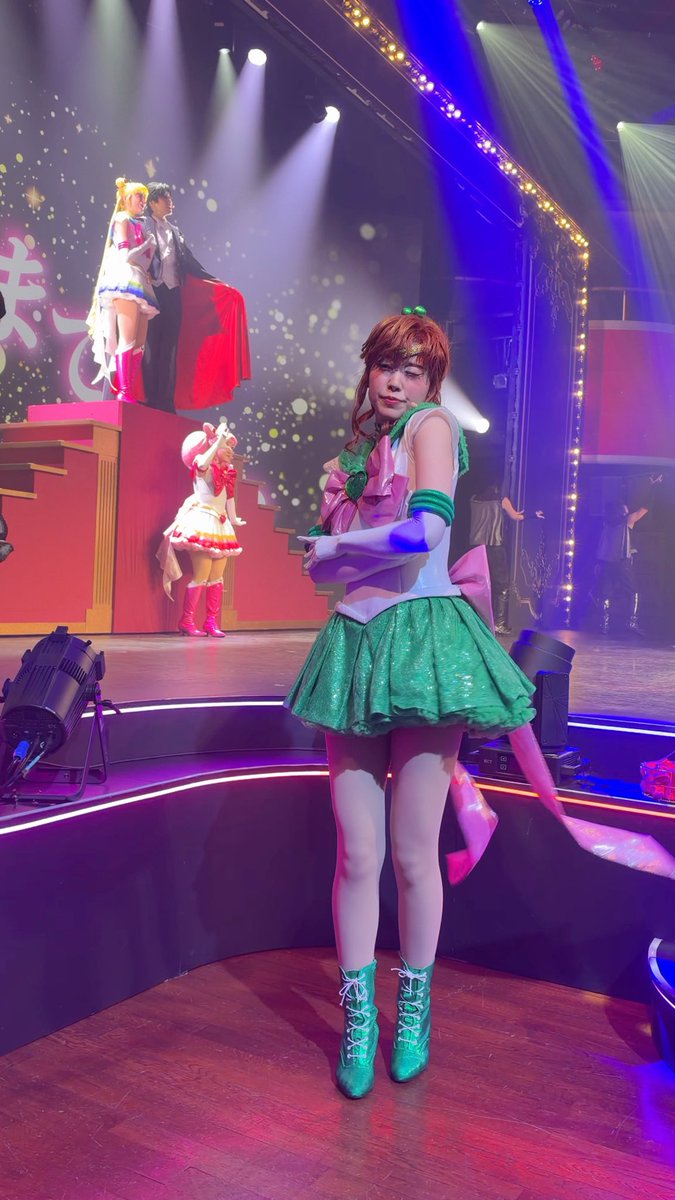 A performer dressed as Sailor Jupiter from Sailor Moon stands on stage in a green and white costume with a green skirt, while other Sailor Moon cosplayers appear on a raised platform behind her.
