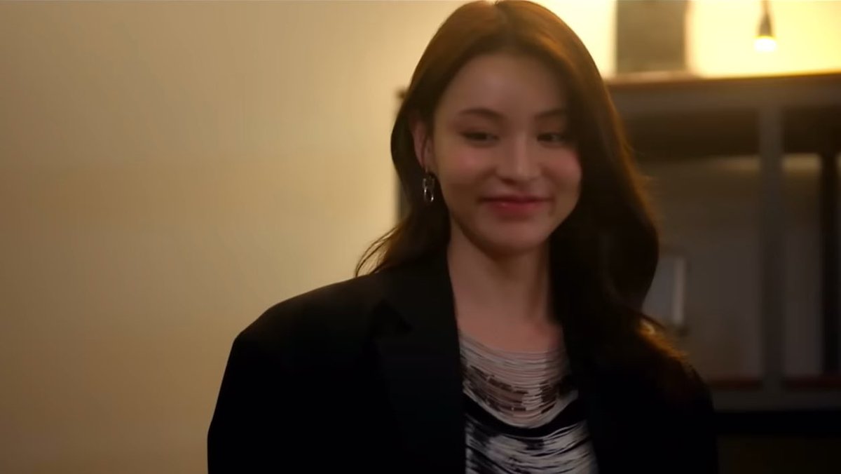 A young woman with long dark hair wearing a black blazer over a patterned top smiles softly in a dimly lit interior setting.