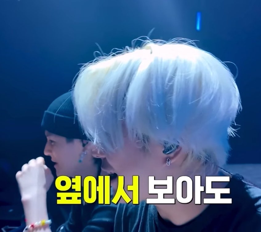 A scene from a Korean variety show or broadcast featuring two young men in dark clothing, one with platinum blonde hair; Korean subtitles in yellow and white read '옆에서 보아도' (Even looking from the side).