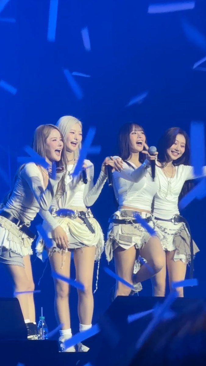 Four female performers in white outfits with ruffled skirts perform on stage under blue lighting as confetti falls around them.