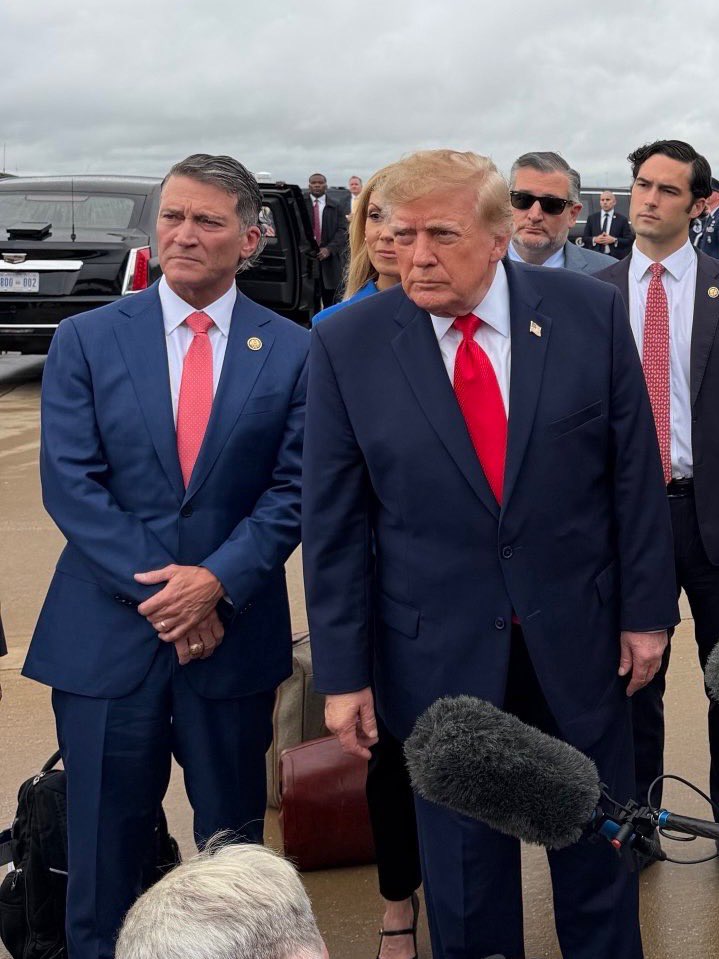 A group of formally dressed people stand outdoors near a black vehicle, with a prominent man in a dark suit and red tie at the center. Several people and recording equipment are visible around him. There is a small amount of text on the vehicle.