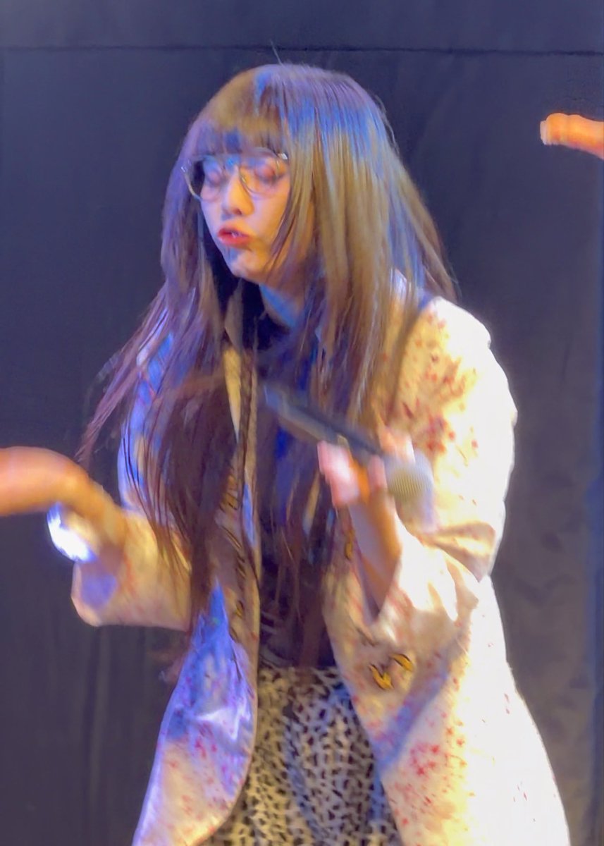 A performer with bangs and glasses, wearing a white jacket with red splatters over a leopard-print top, is captured in motion on a dimly lit stage.
