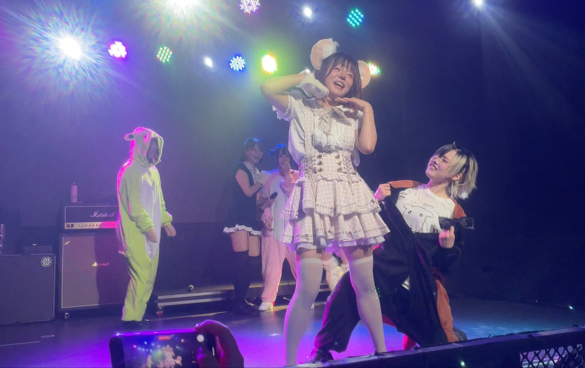 A live performance on a brightly lit stage features performers in costumes, including a person in a green animal onesie and a woman in a white frilly dress with mouse ears posing playfully for the audience.